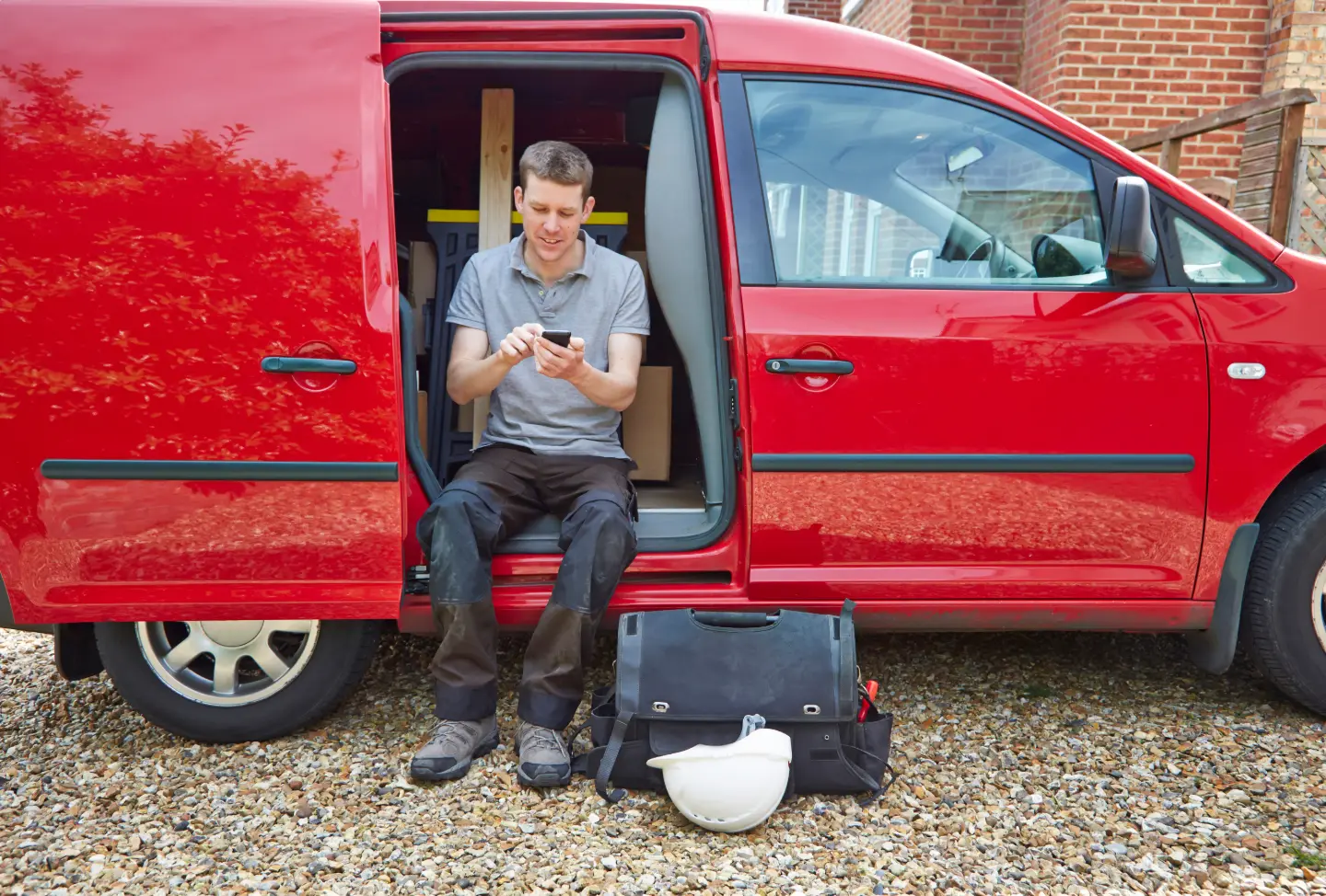 A tradesman sitting in the open side door of his red work van, checking an estimate on his phone